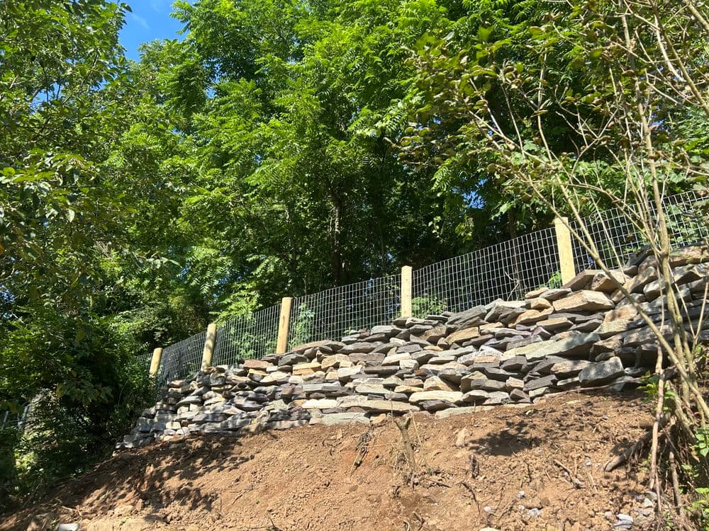 Retaining wall made of stones with wooden fence and greenery above in a sunny landscape.