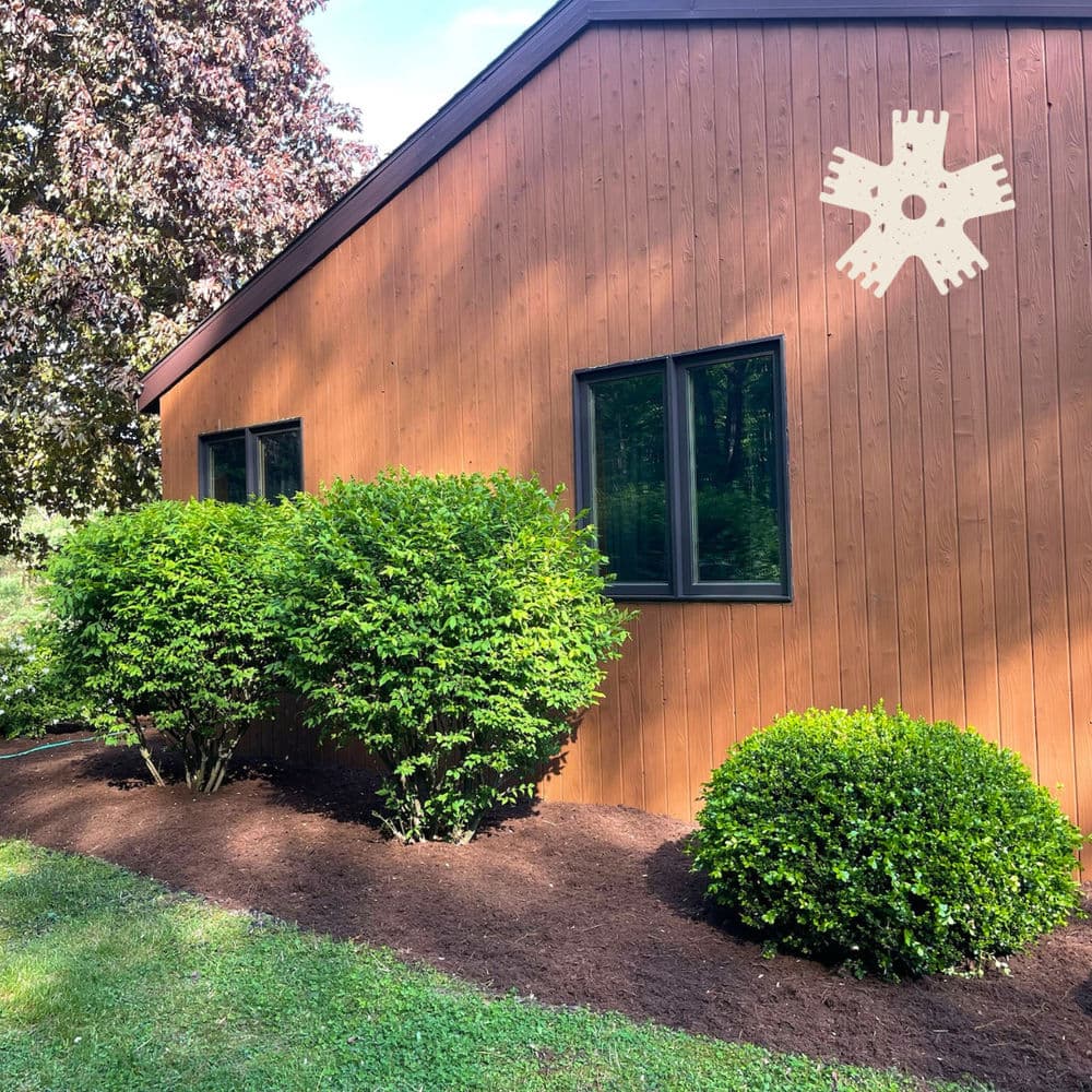 Brown house exterior with large windows, surrounded by lush green bushes and fresh mulch.